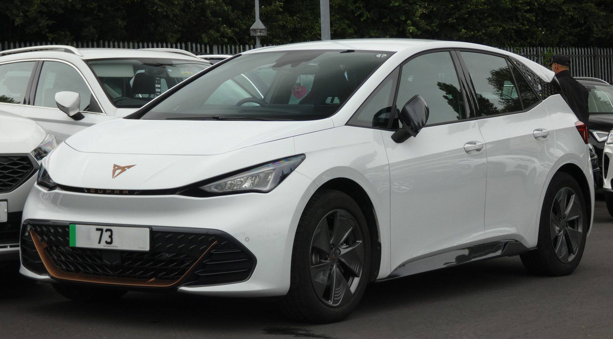 A white Cupra Born on UK dealer plates parked among other cars, front three-quarter view