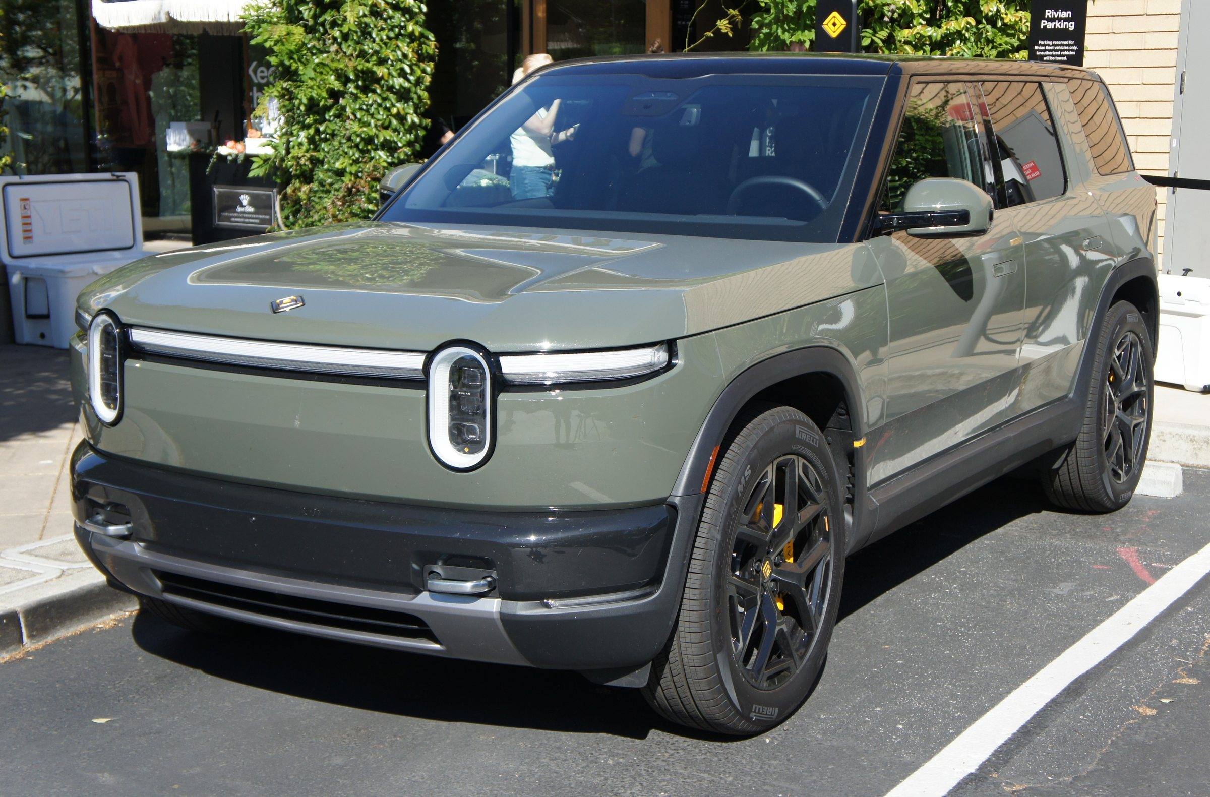 A green Rivian R2 parked at a kerb outside a shop, front three-quarter view
