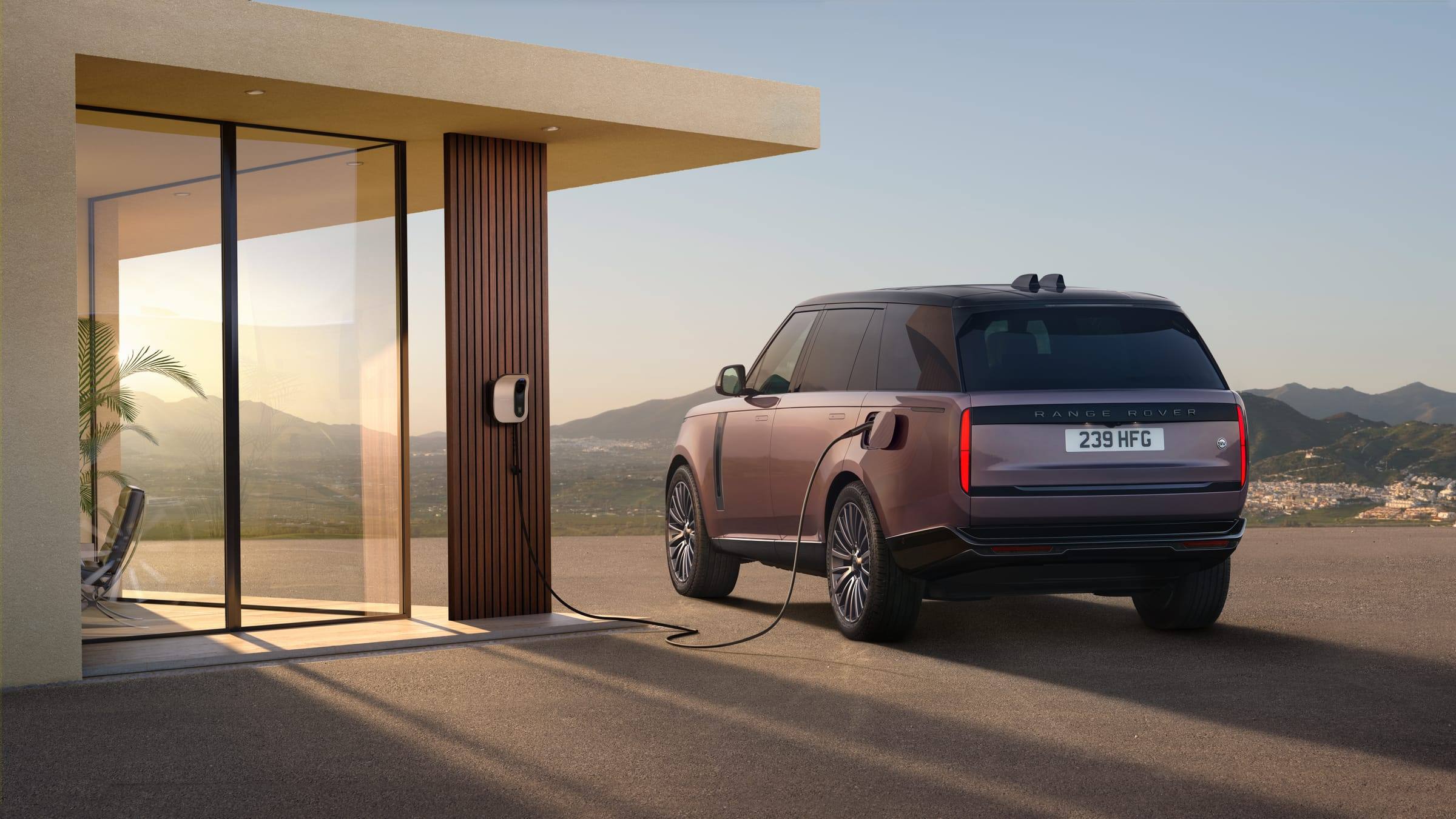 A Range Rover Electric plugged into a wall-mounted charger outside a modern house at sunset, seen from the rear three-quarter