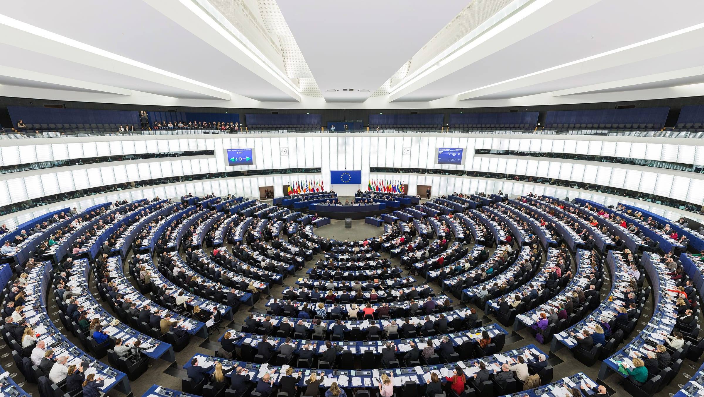 The European Parliament's semicircular plenary chamber in Strasbourg, seen from the back, with members at their desks and EU flags behind the presidency