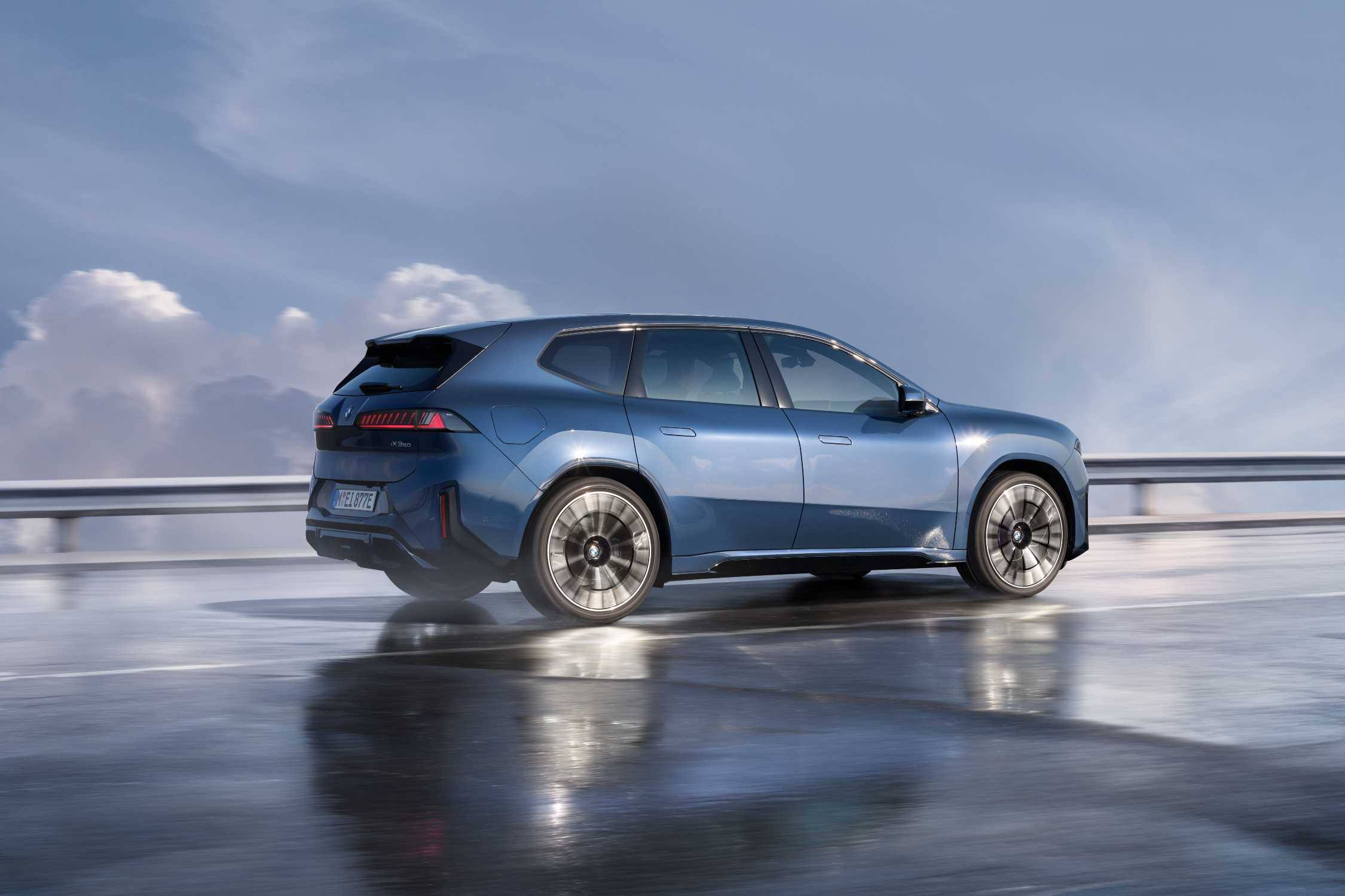 A blue BMW iX3 driving on a wet road with a crash barrier and clouds behind, rear three-quarter view