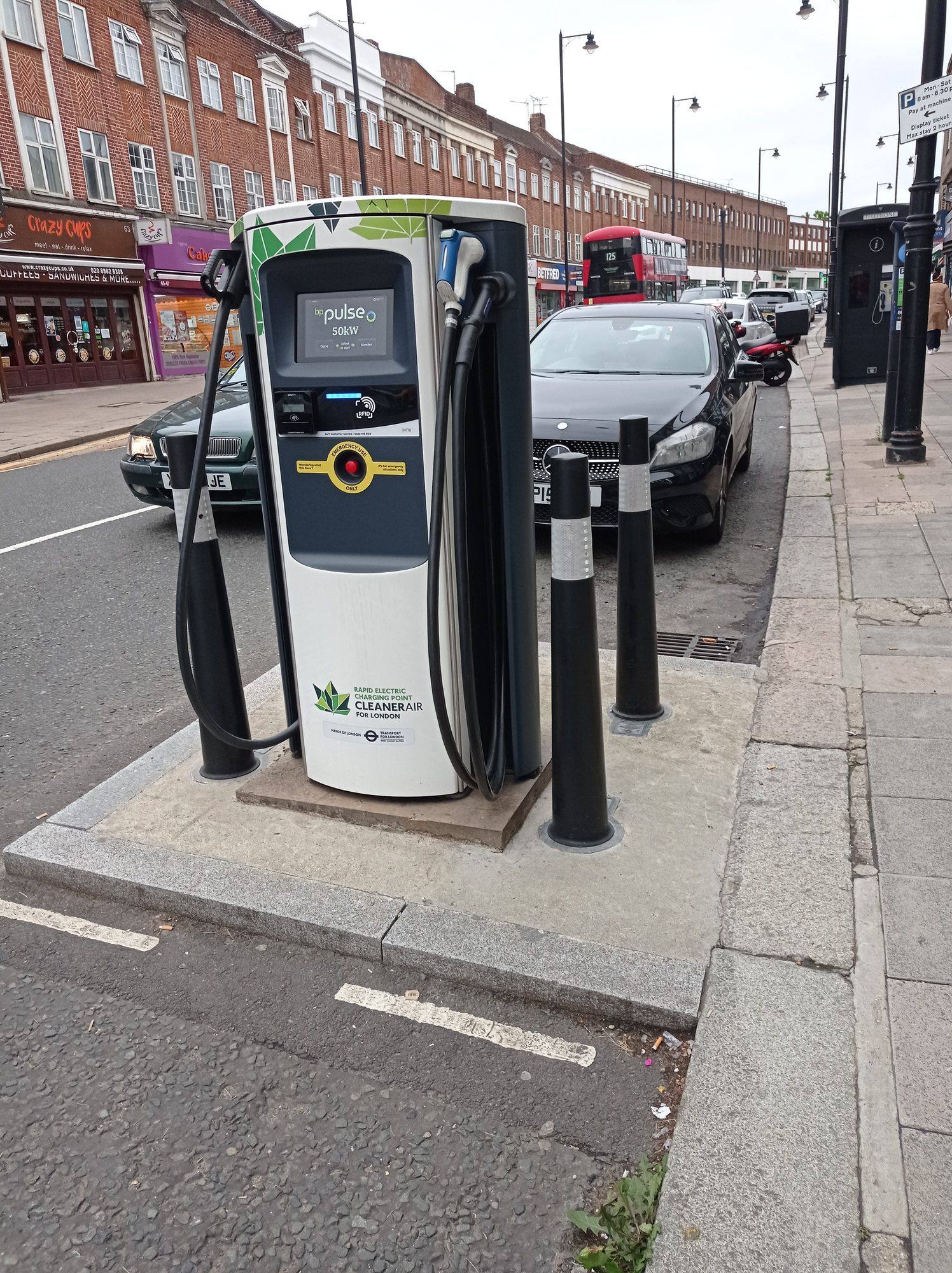 A white BP Pulse 50 kW charger on a kerbside island in a London high street, with parked cars and a red double-decker bus behind