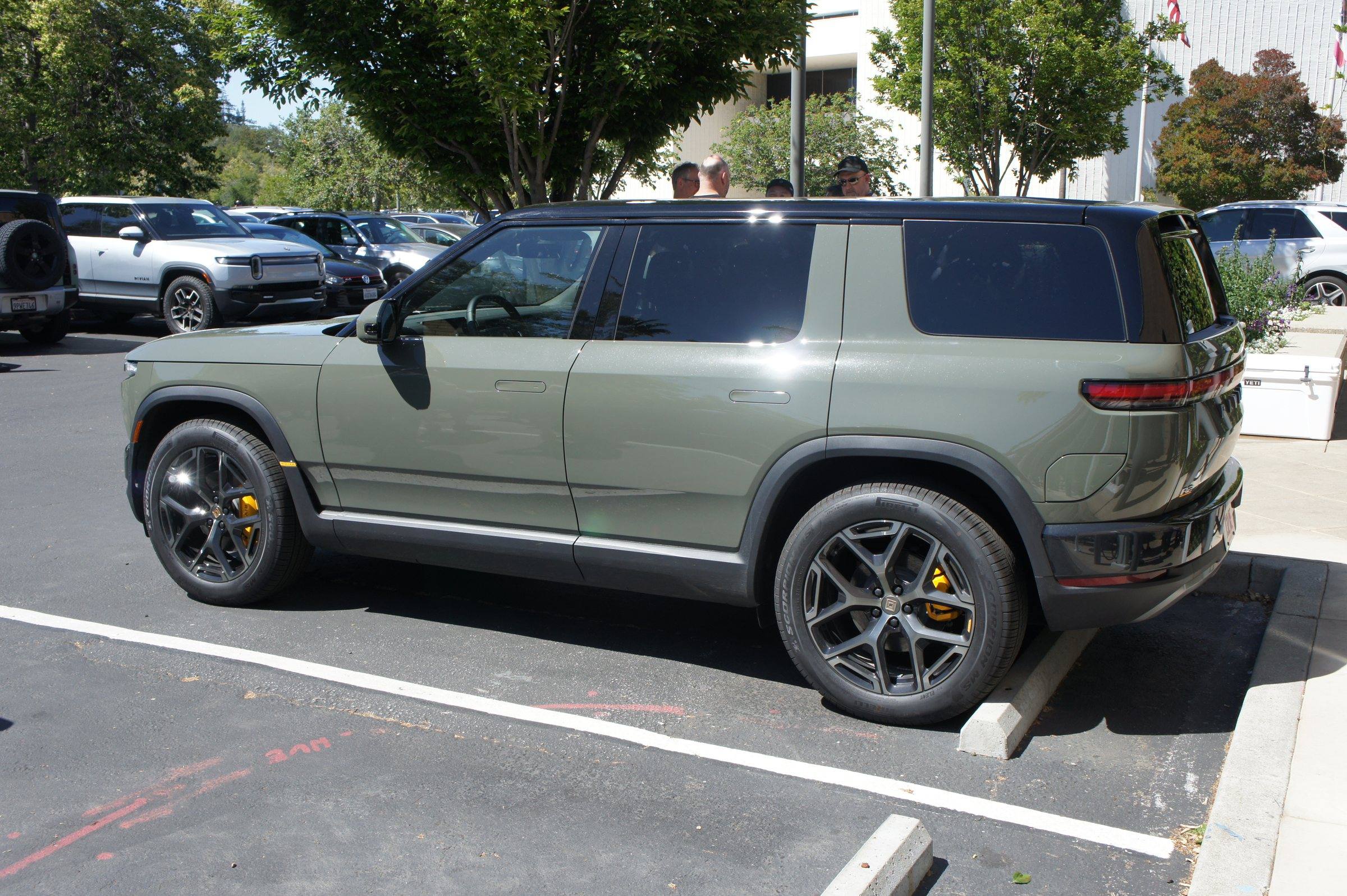 A green Rivian R2 parked in a car park, side and rear view, showing its dark multi-spoke wheels