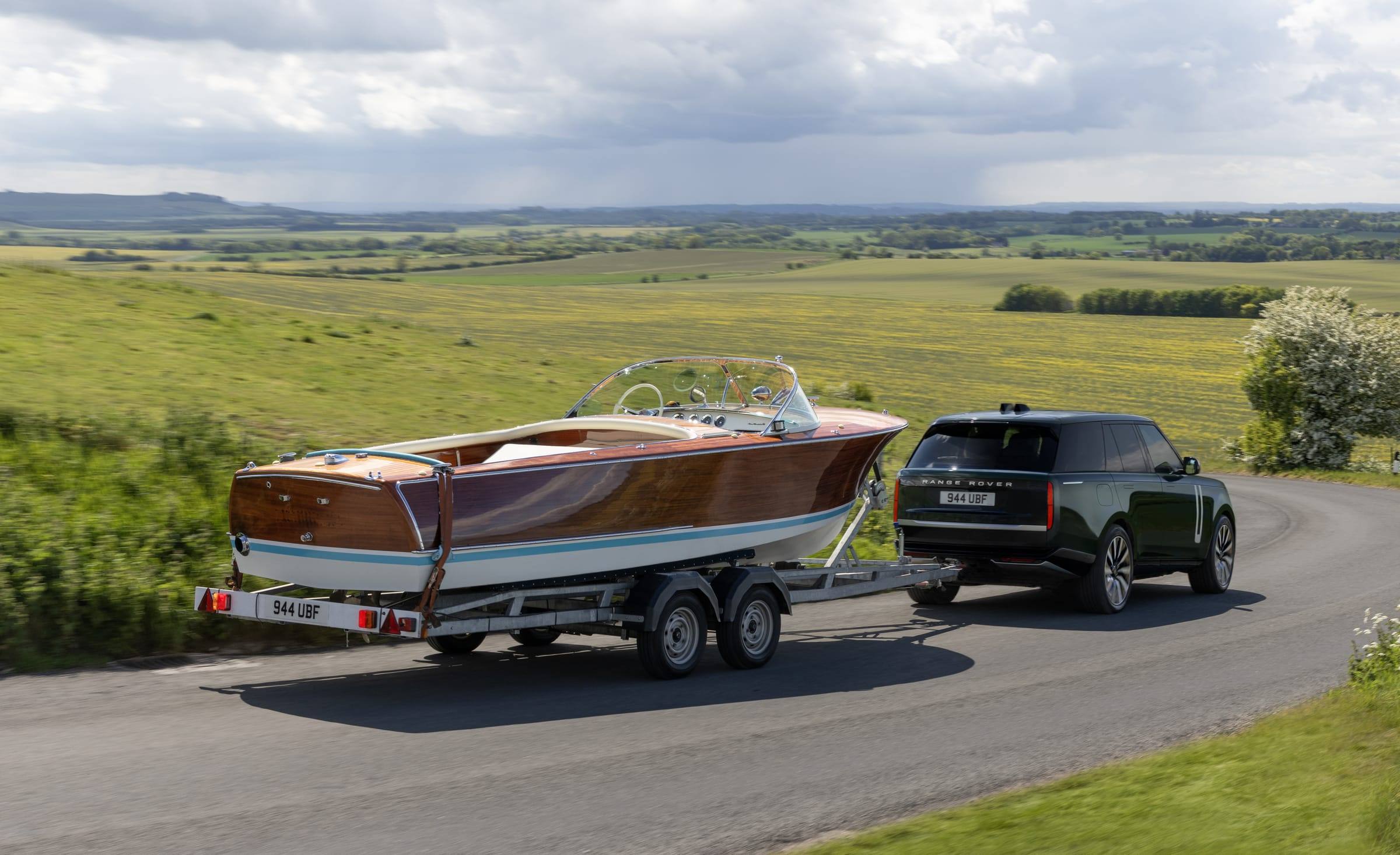 A dark Range Rover Electric towing a wooden speedboat on a twin-axle trailer along a country road