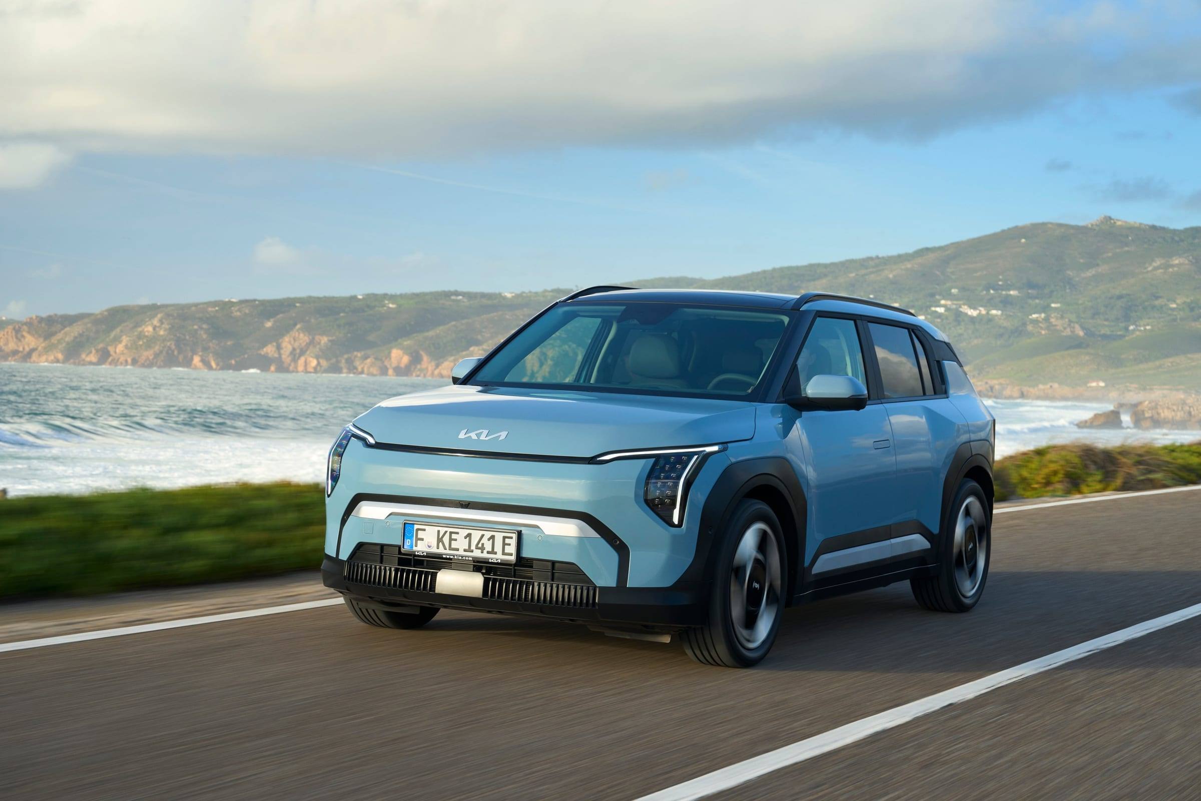 A light blue Kia EV3 driving along a coastal road, seen from the front three-quarter view