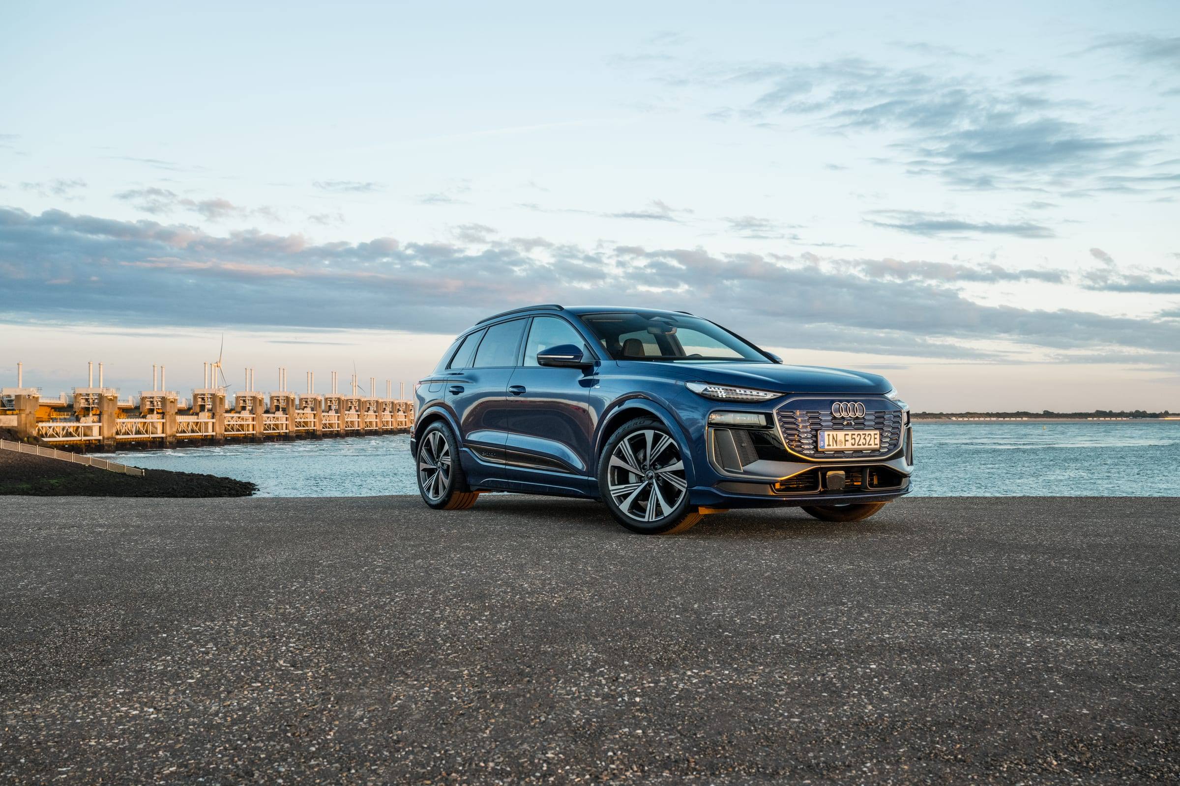 A blue Audi Q6 e-tron parked on a quayside by the sea at dusk, seen from the front three-quarter view