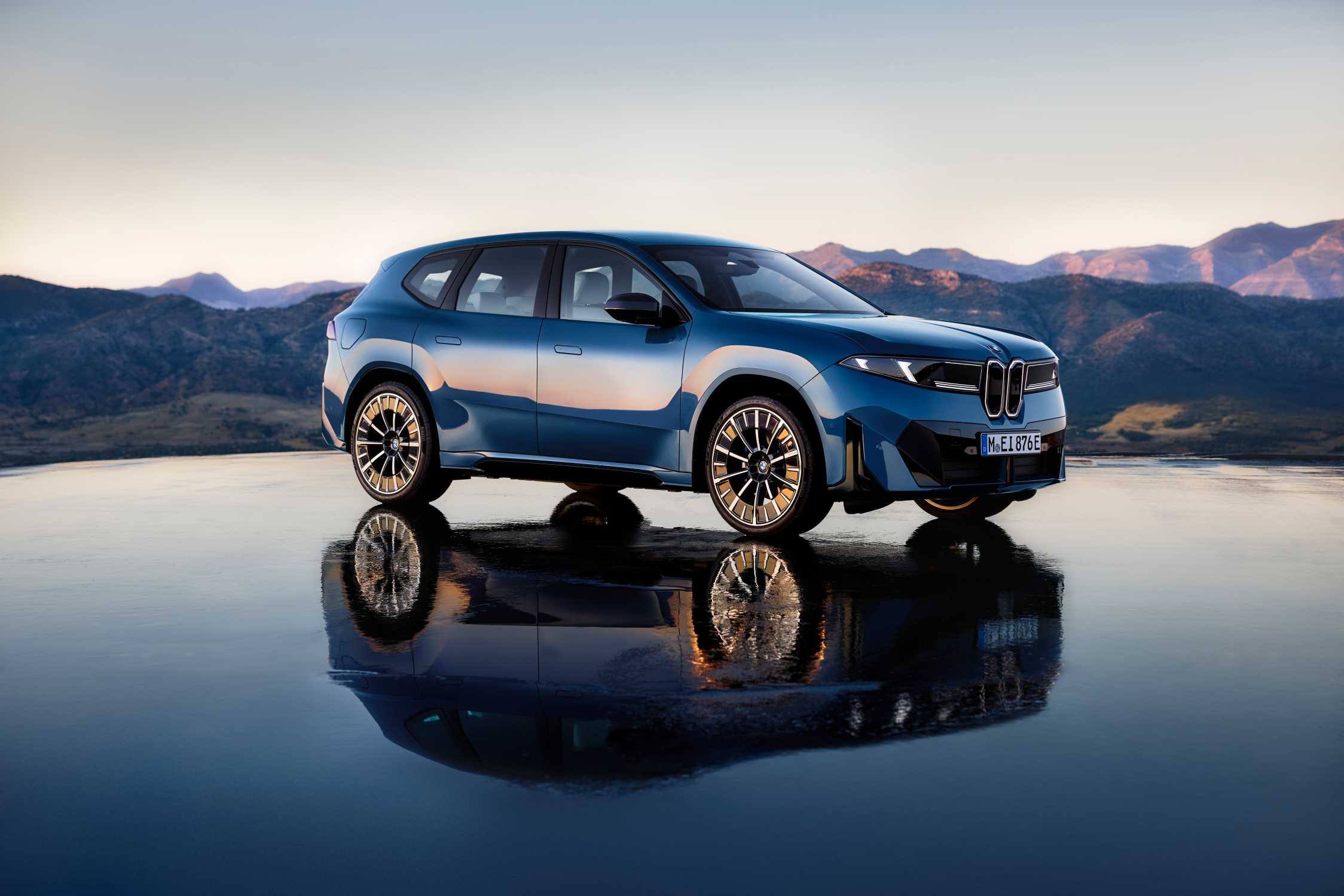 A blue BMW iX3 parked on a wet, reflective surface with hills behind it at dusk, seen from the front three-quarter