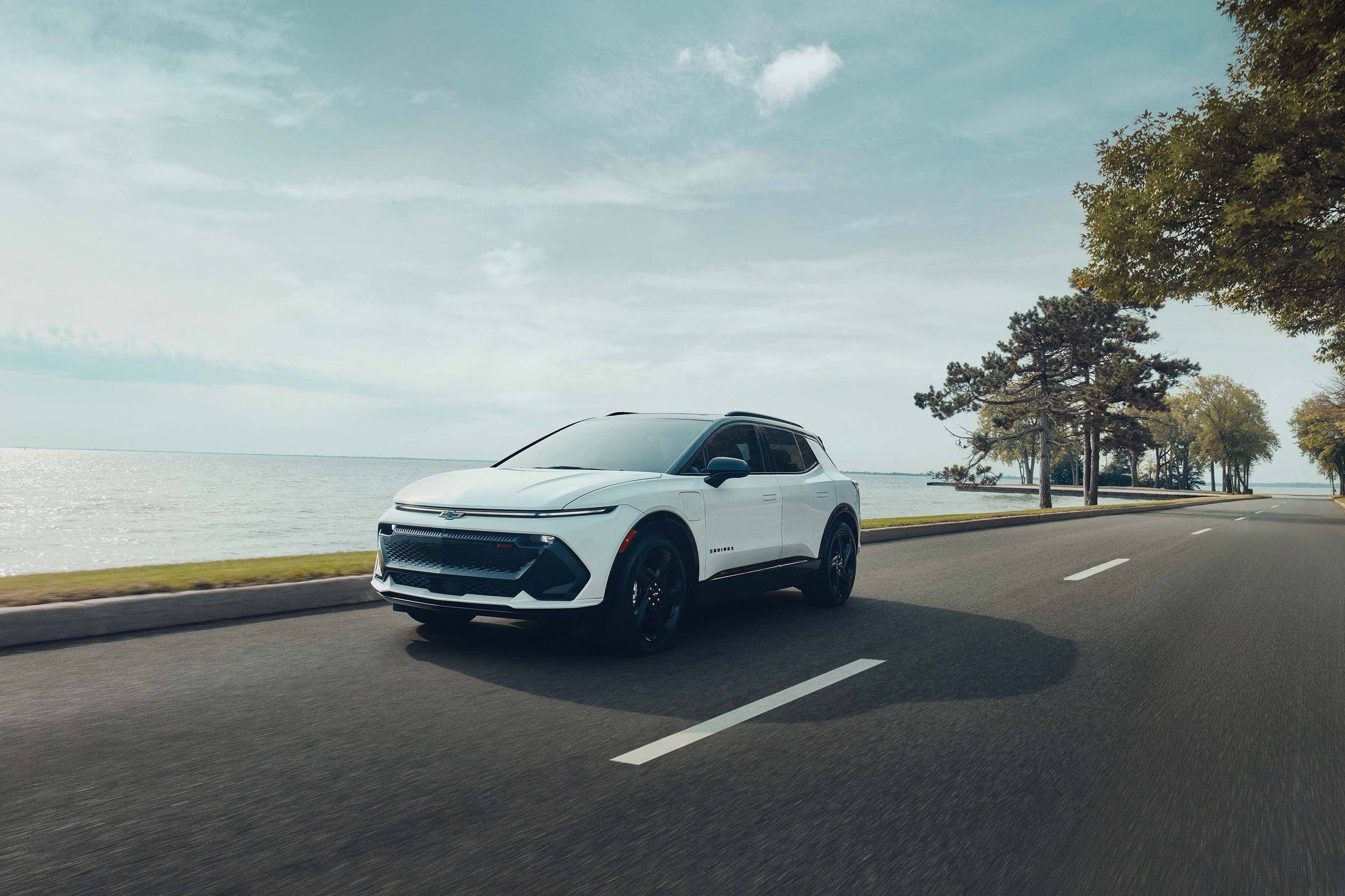 White Chevrolet Equinox EV RS, front three-quarter view, driving on a lakeside road