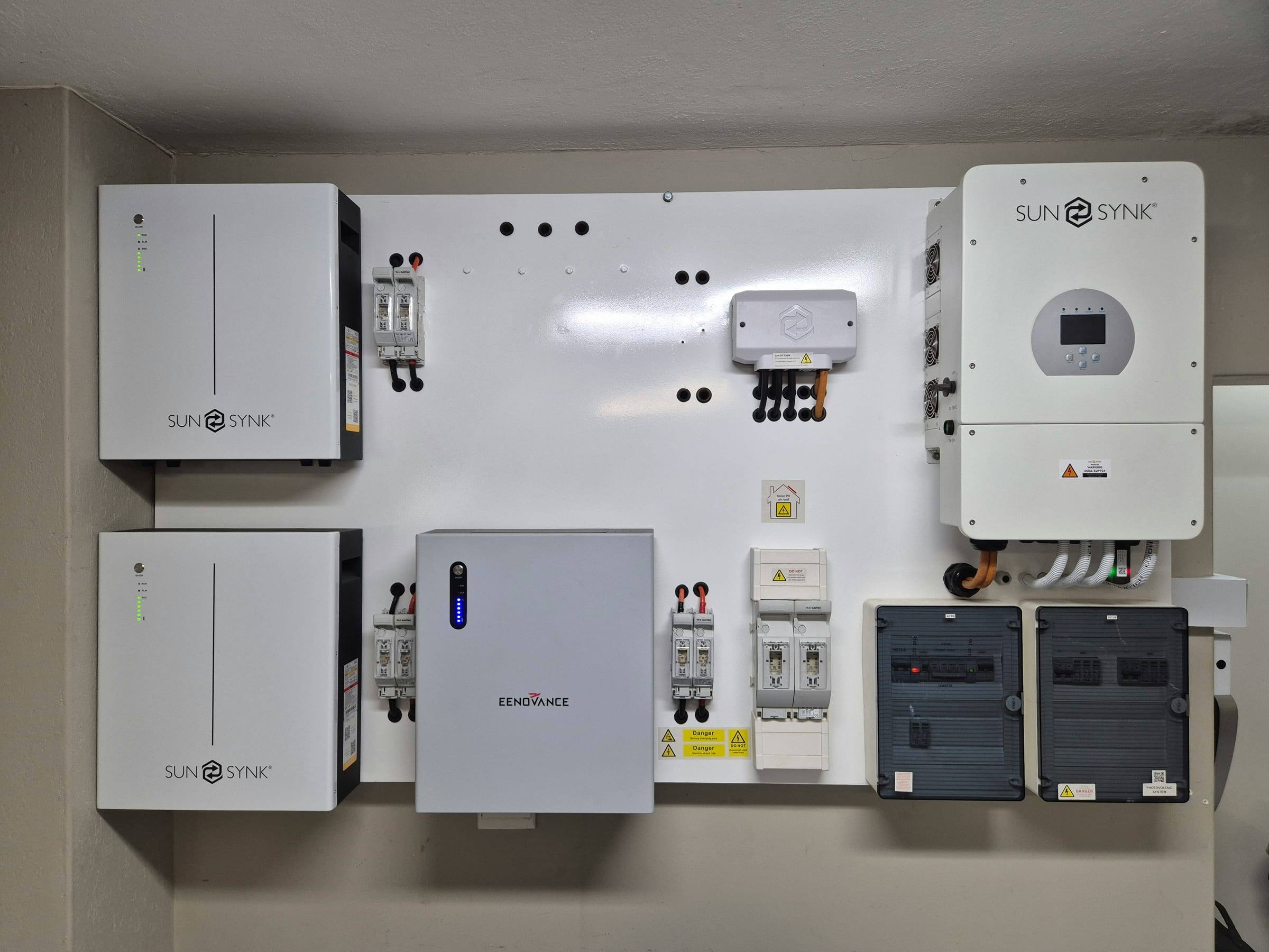 A wall-mounted home energy system with three battery cabinets, a solar inverter and two breaker boxes on a white backboard