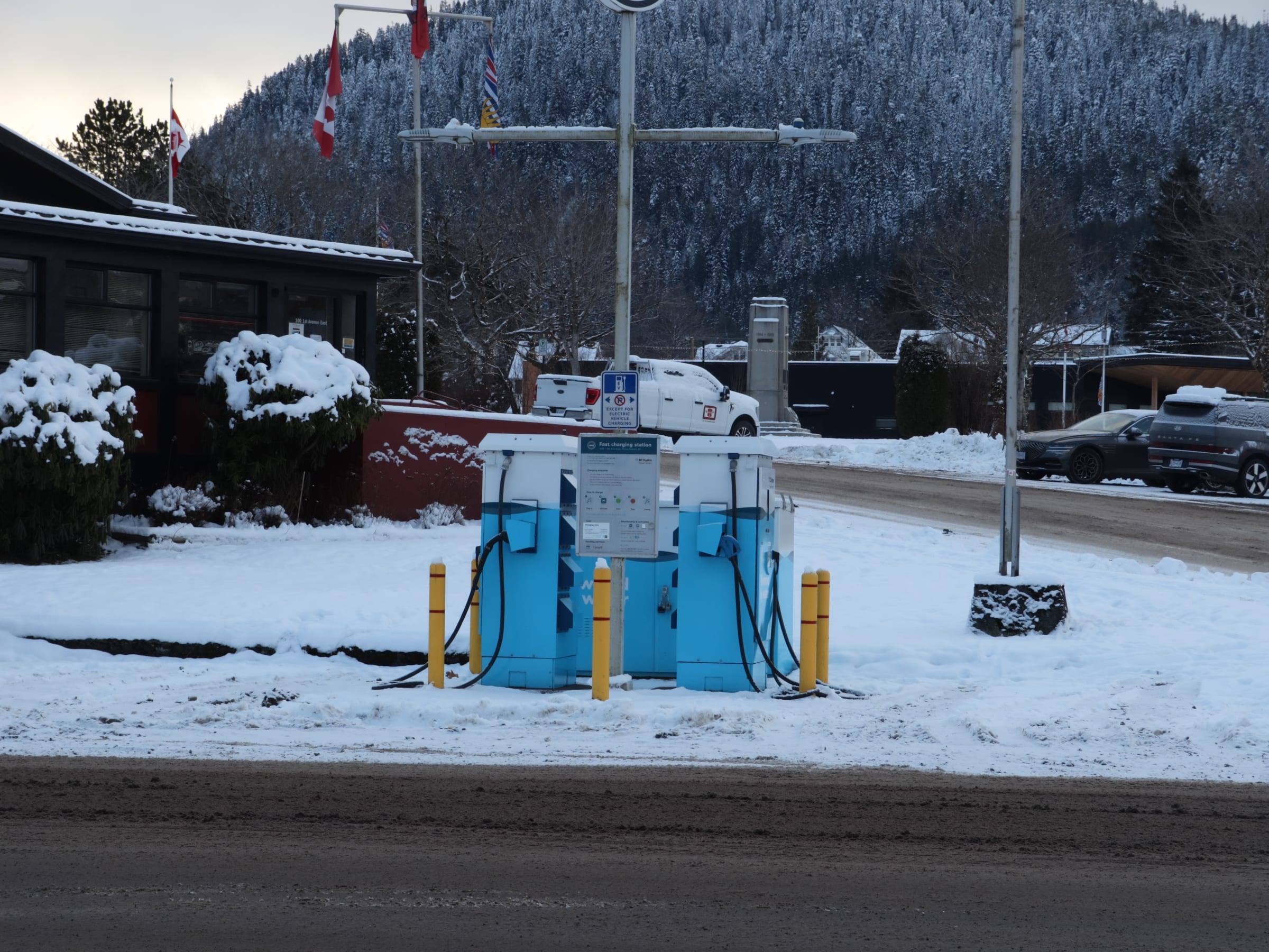 Blue public fast chargers standing in fresh snow beside a road, with snow-covered hills behind