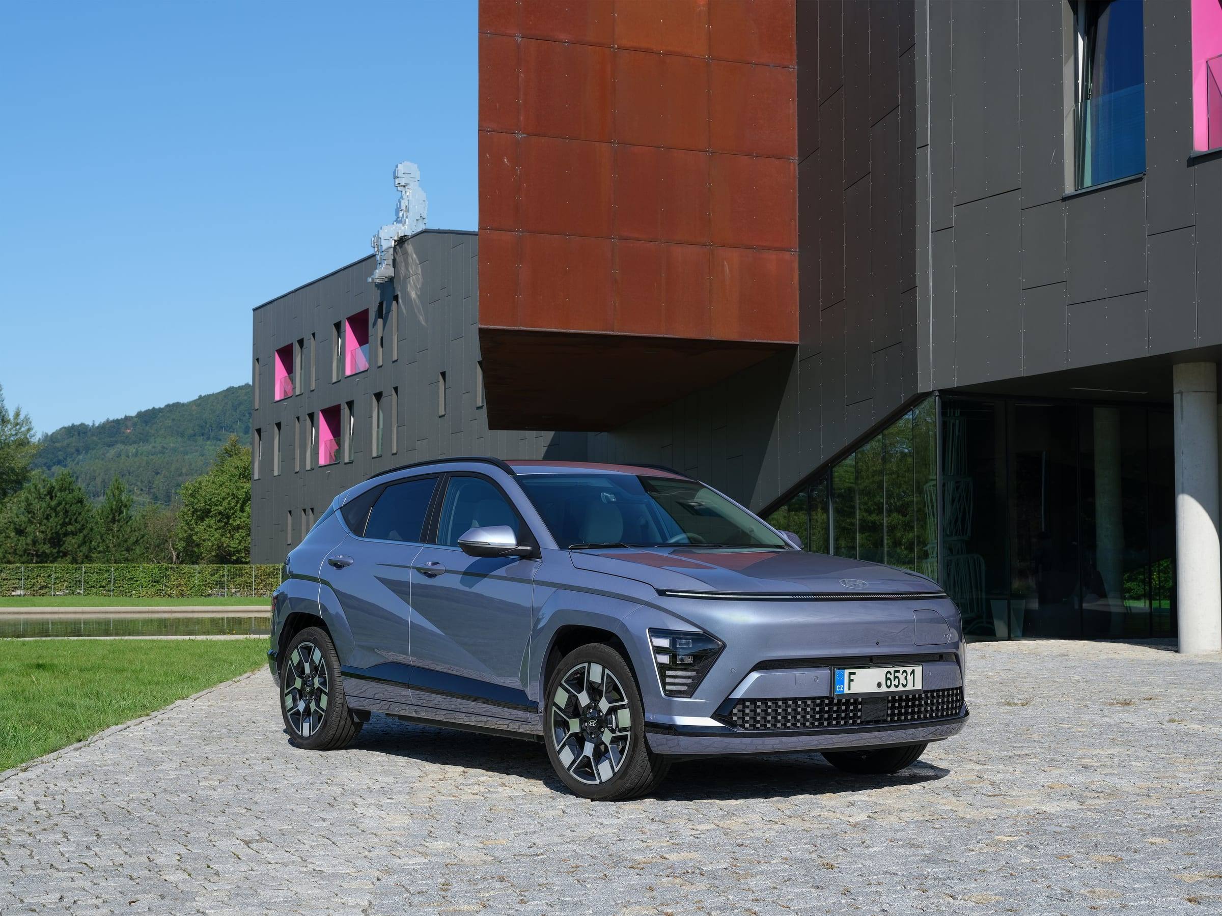 Blue-grey Hyundai Kona Electric, front three-quarter view, parked on cobbles in front of a modern building