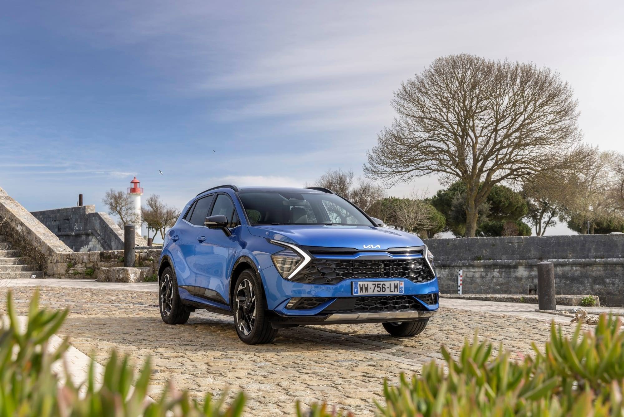 Blue Kia Sportage Plug-in Hybrid (2022 model, before the facelift), front three-quarter view parked on cobbles beside a stone wall