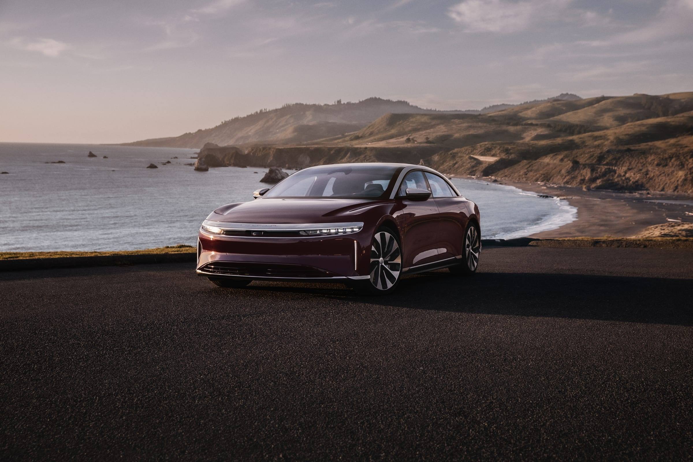 Dark red Lucid Air Grand Touring, front three-quarter view, parked above a coastline