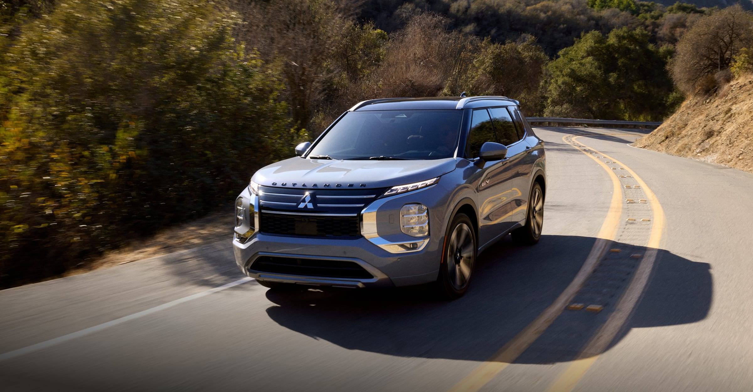 A silver 2026 Mitsubishi Outlander PHEV driving along a winding hillside road, seen from the front three-quarter view