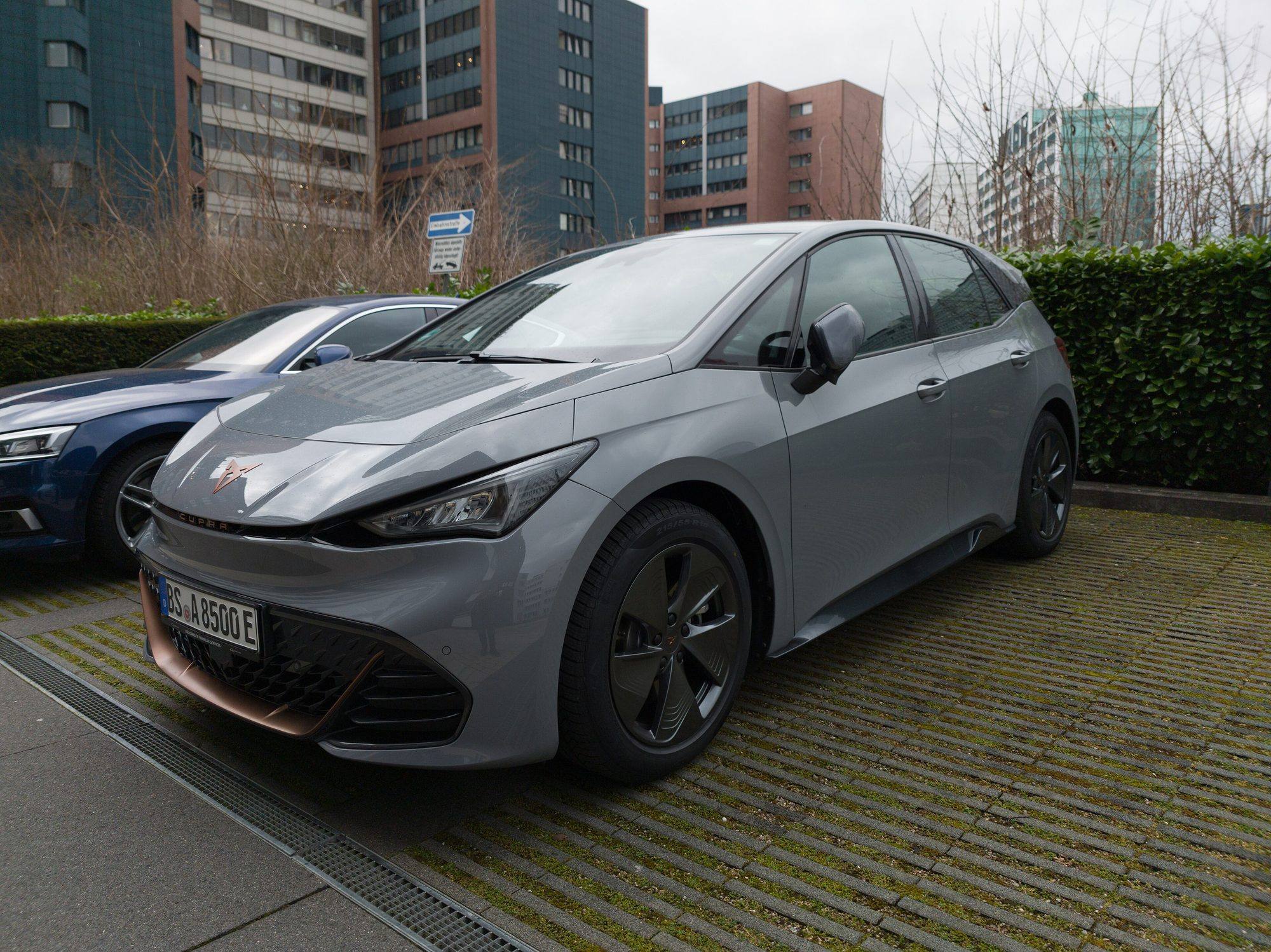A grey Cupra Born hatchback with copper trim parked on a grass-paver car park in front of office blocks