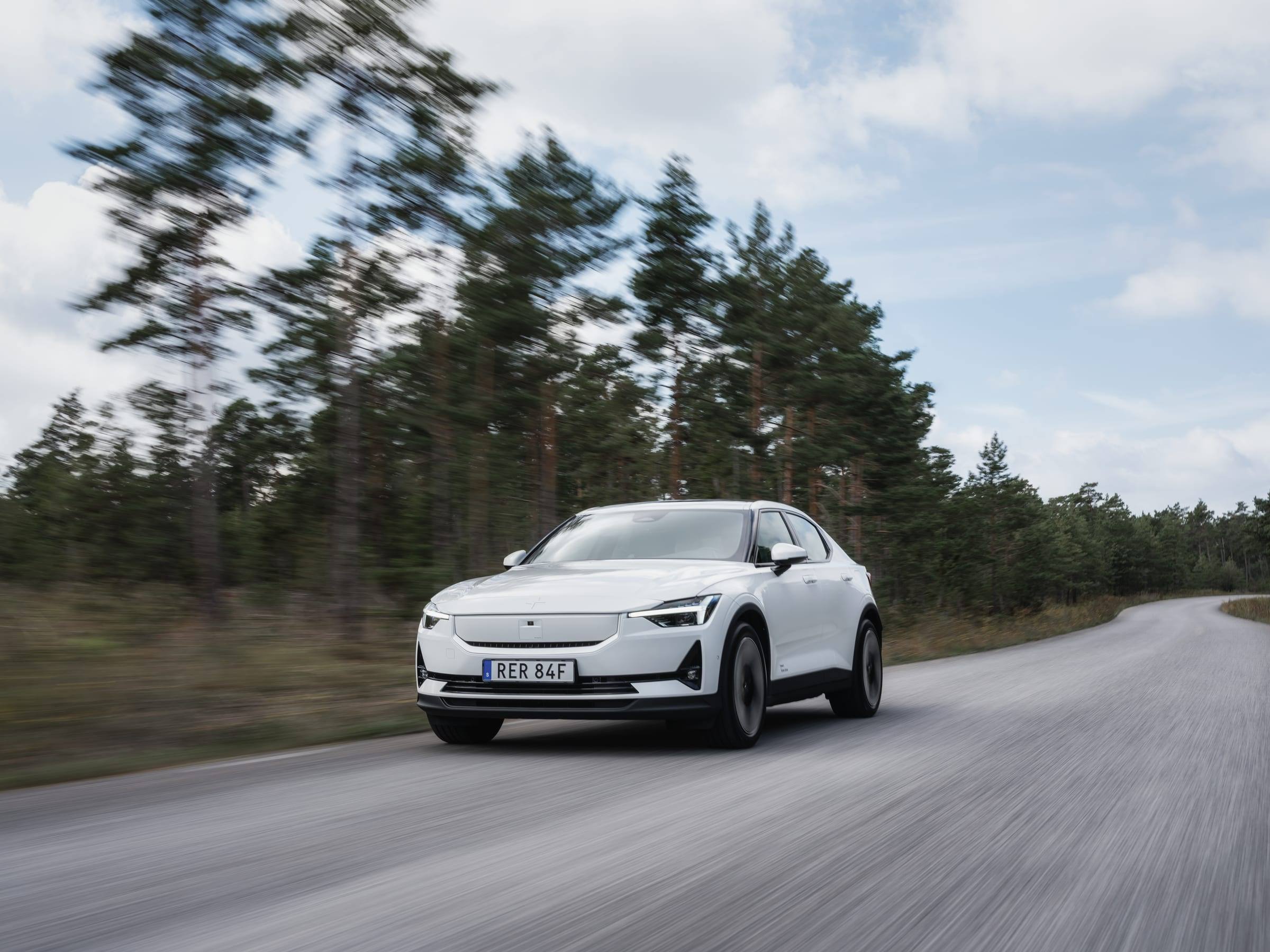 A white Polestar 2 driving on a country road through pine woods, seen from the front three-quarter view