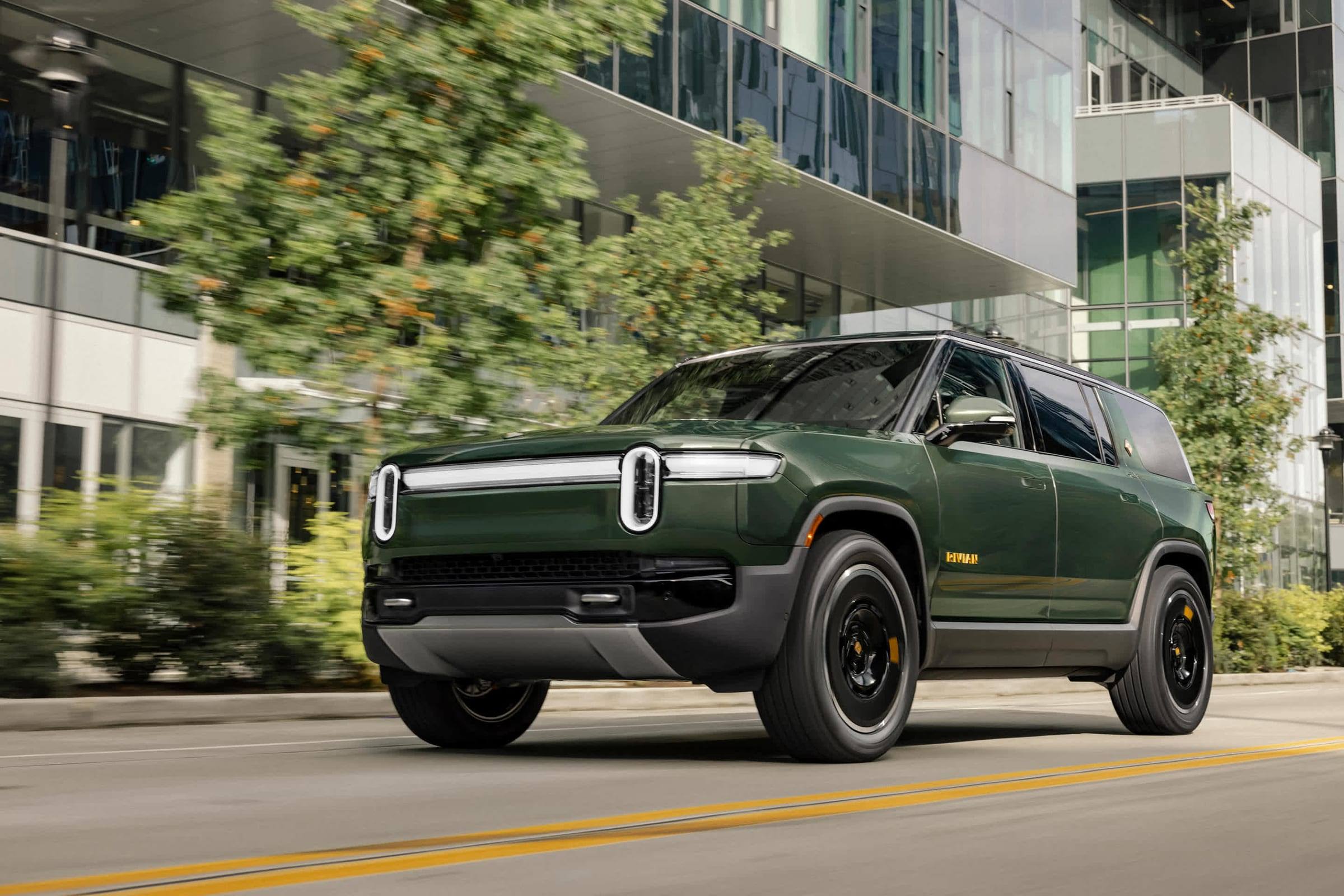 Dark green Rivian R1S, front three-quarter view, driving past modern glass buildings