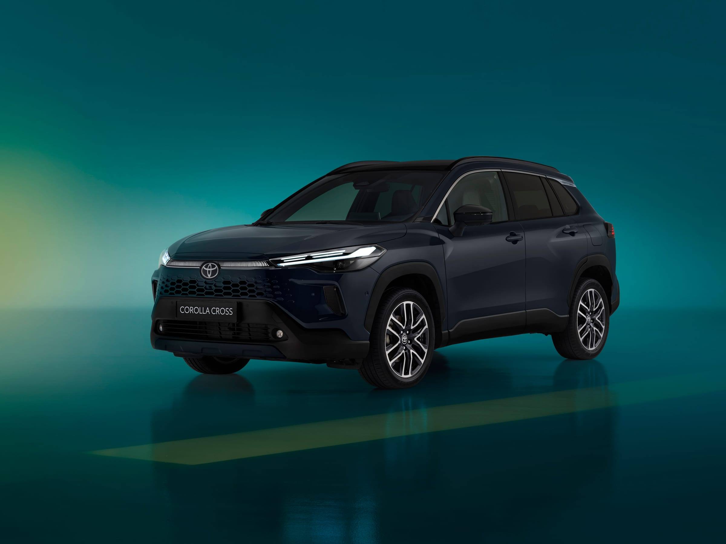 A dark blue updated Toyota Corolla Cross in a teal-lit studio, seen from the front three-quarter view