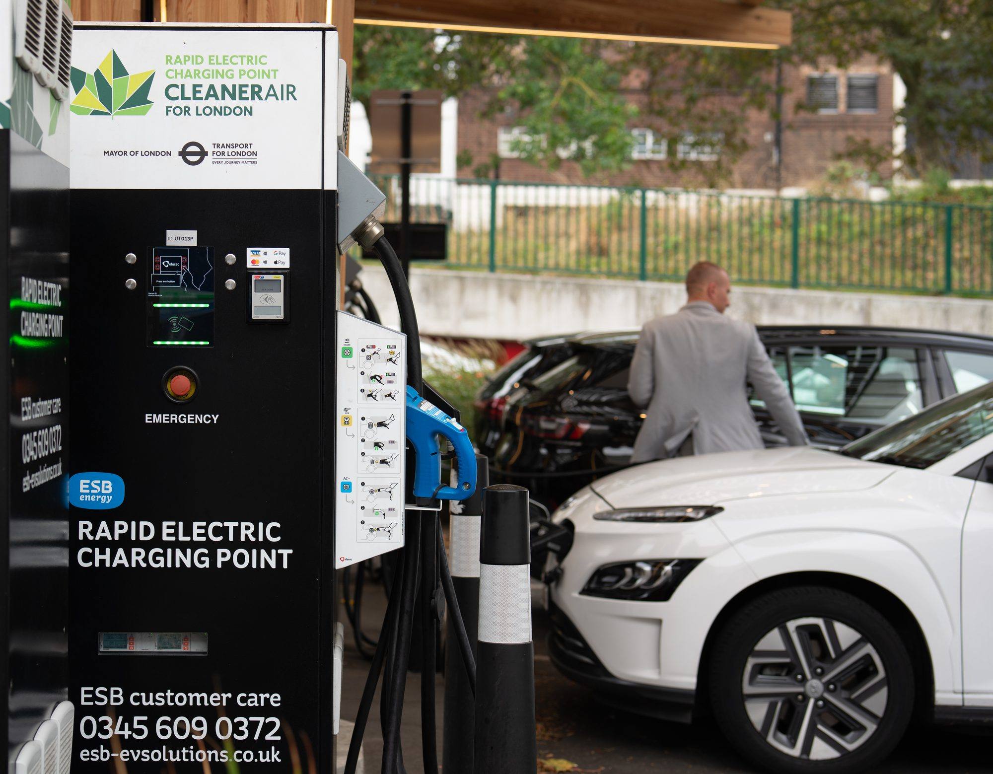 A black ESB Energy rapid charging point with a blue connector, next to a white electric car plugged in under a timber canopy in London