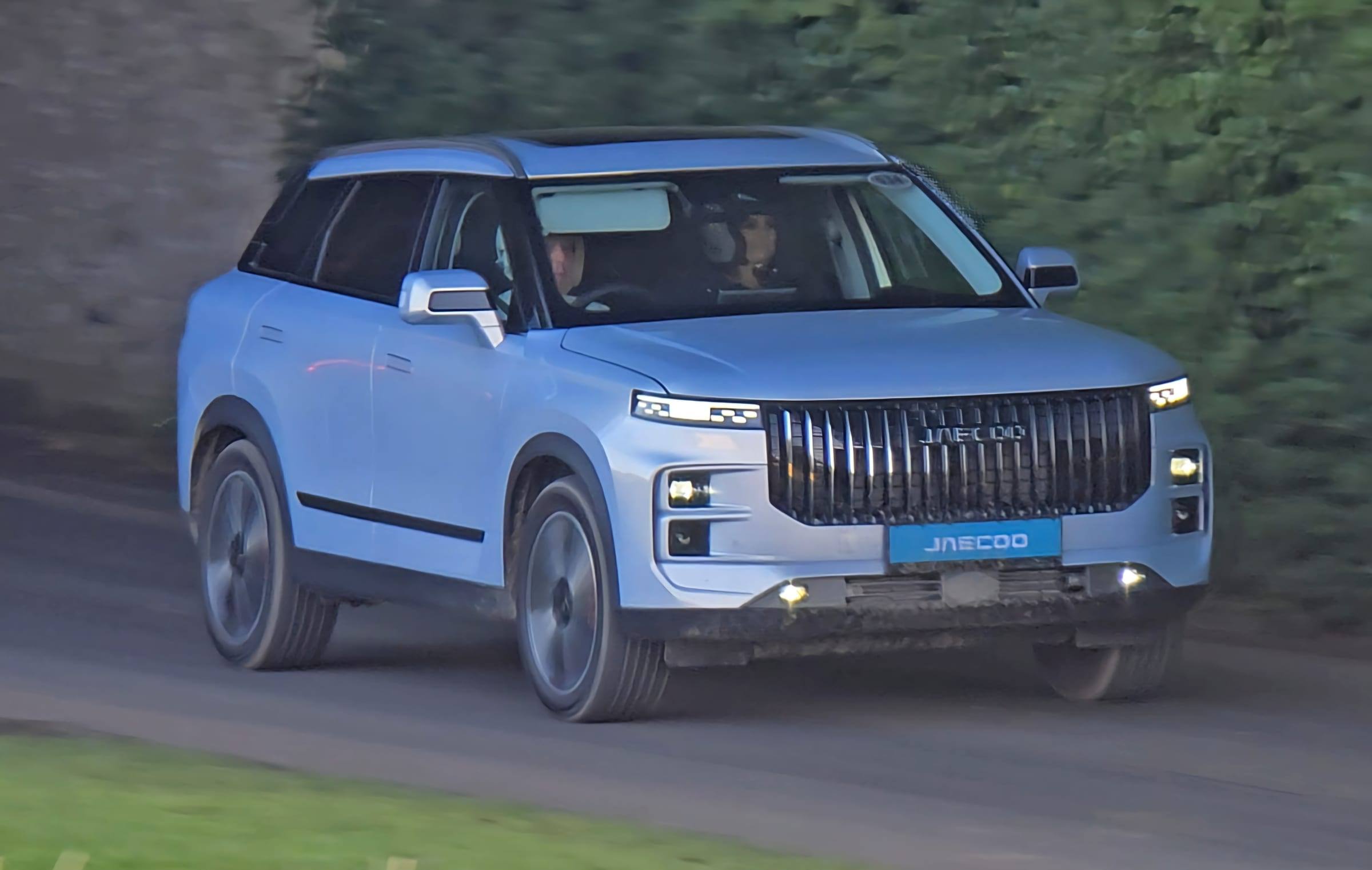 A pale blue Jaecoo 7 SUV driving along a tree-lined track