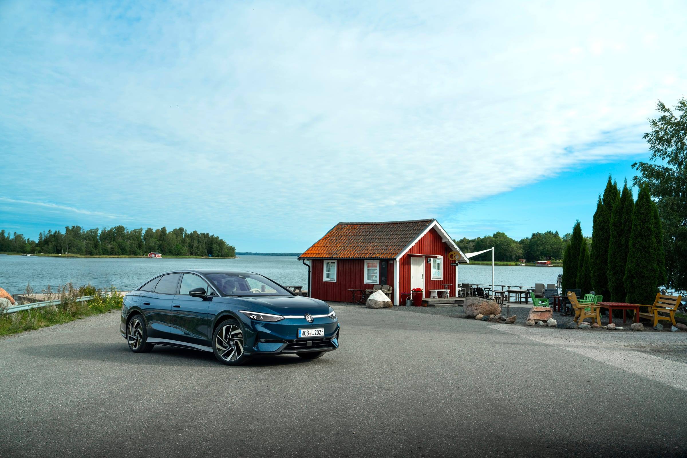 Dark blue Volkswagen ID.7 Pro S, front three-quarter view, parked by a lake next to a red wooden house
