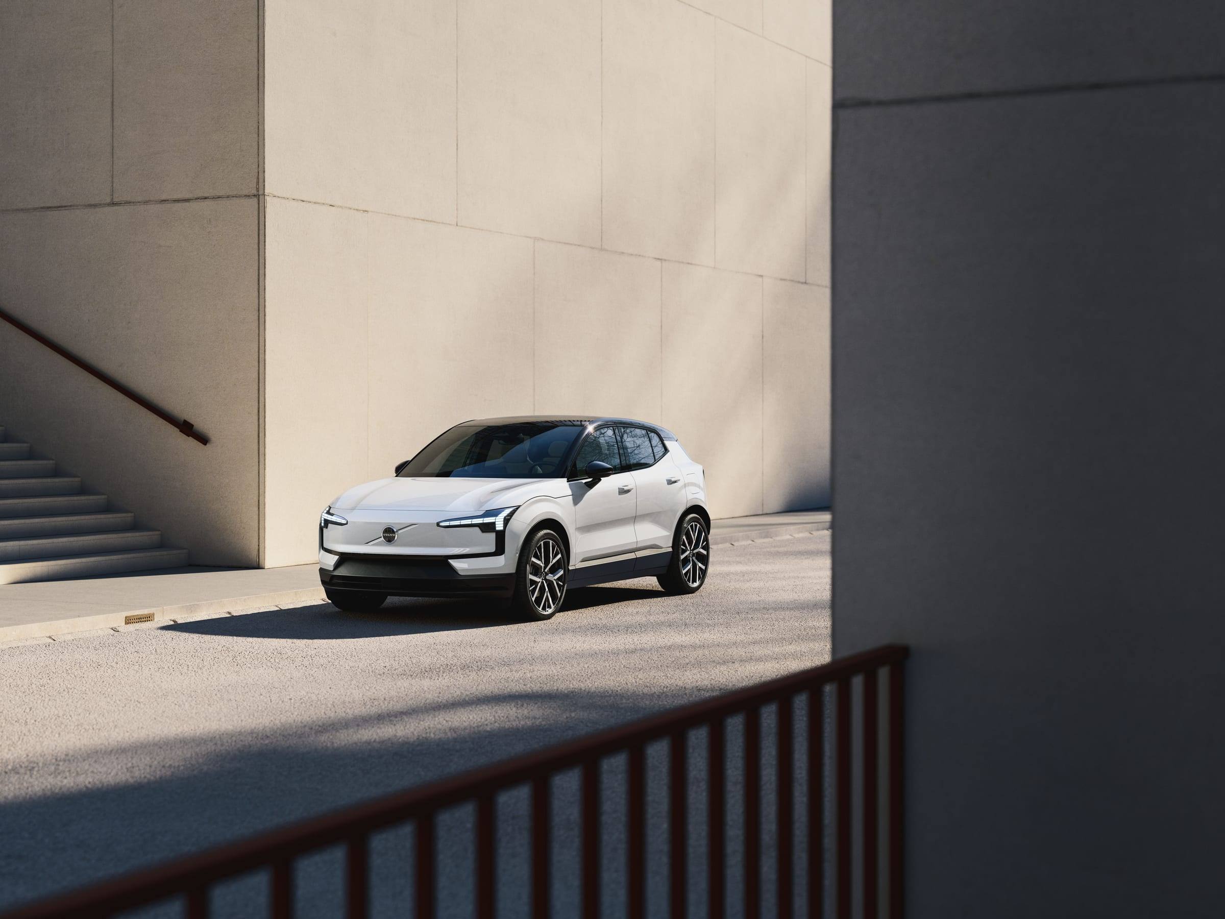 A white Volvo EX30 parked on a street beside tall concrete walls, seen from the front three-quarter view